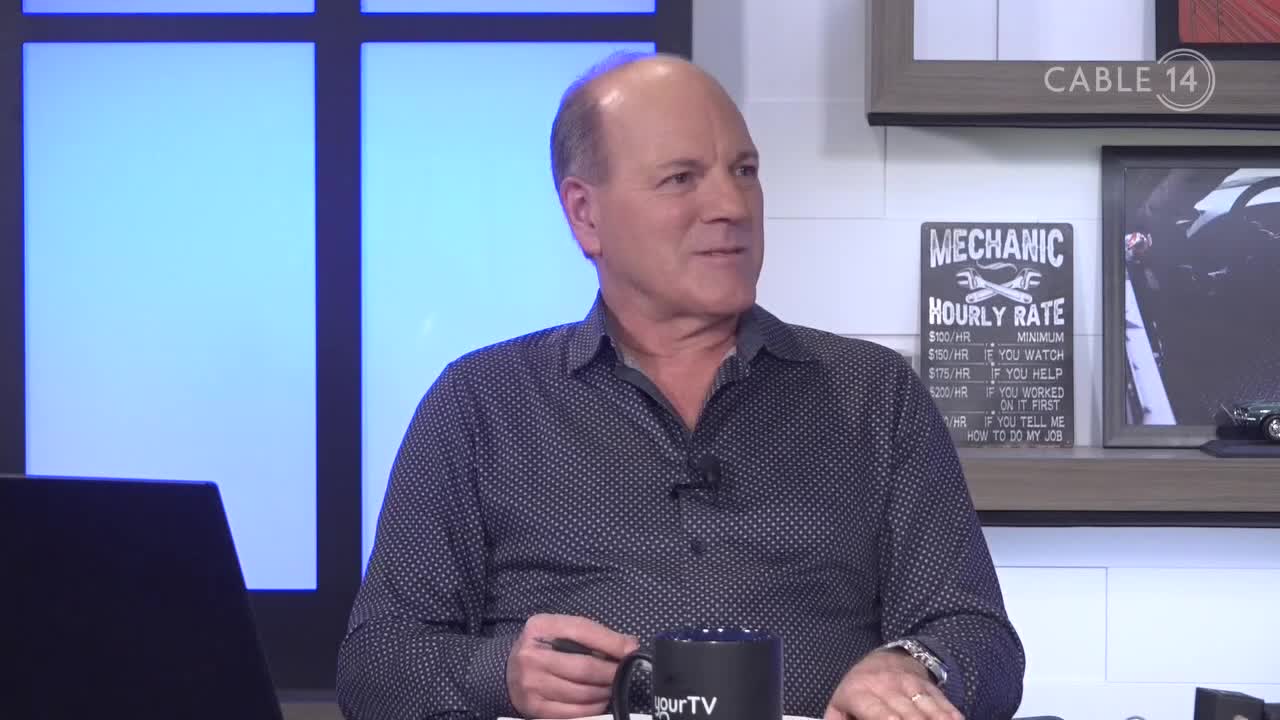 A man in a patterned shirt sits at a desk, a mug in front of him. The Cable 14 logo is visible, and a "Mechanic Hourly Rate" sign hangs on the wall behind him.
