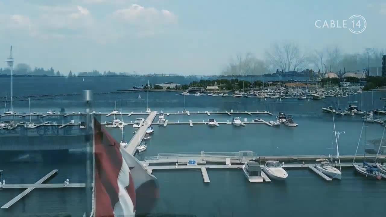 Boats bob gently in the harbor, their masts reaching toward the sky. The Canadian flag waves in the foreground, with Cable 14's logo visible in the upper right.
