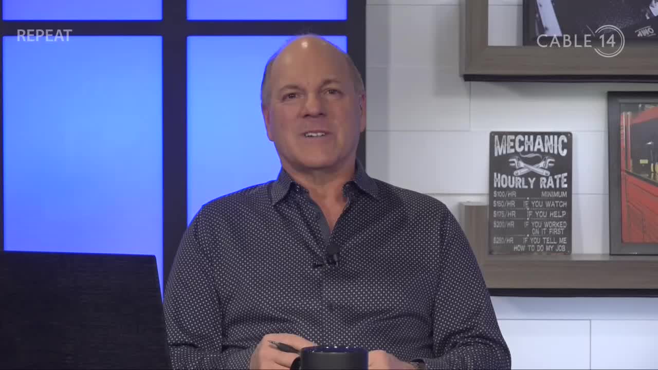 He's speaking directly to the camera, a Cable 14 Hamilton backdrop behind him. A "Mechanic Hourly Rate" sign hangs on the wall to his right.
