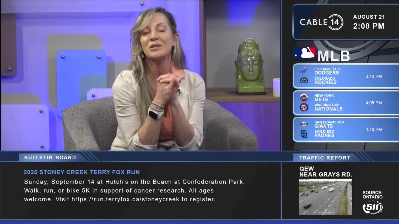 A woman with blonde hair sits in a chair, speaking on Cable 14 Hamilton. The MLB scores and a traffic report flash on the screen beside her.
