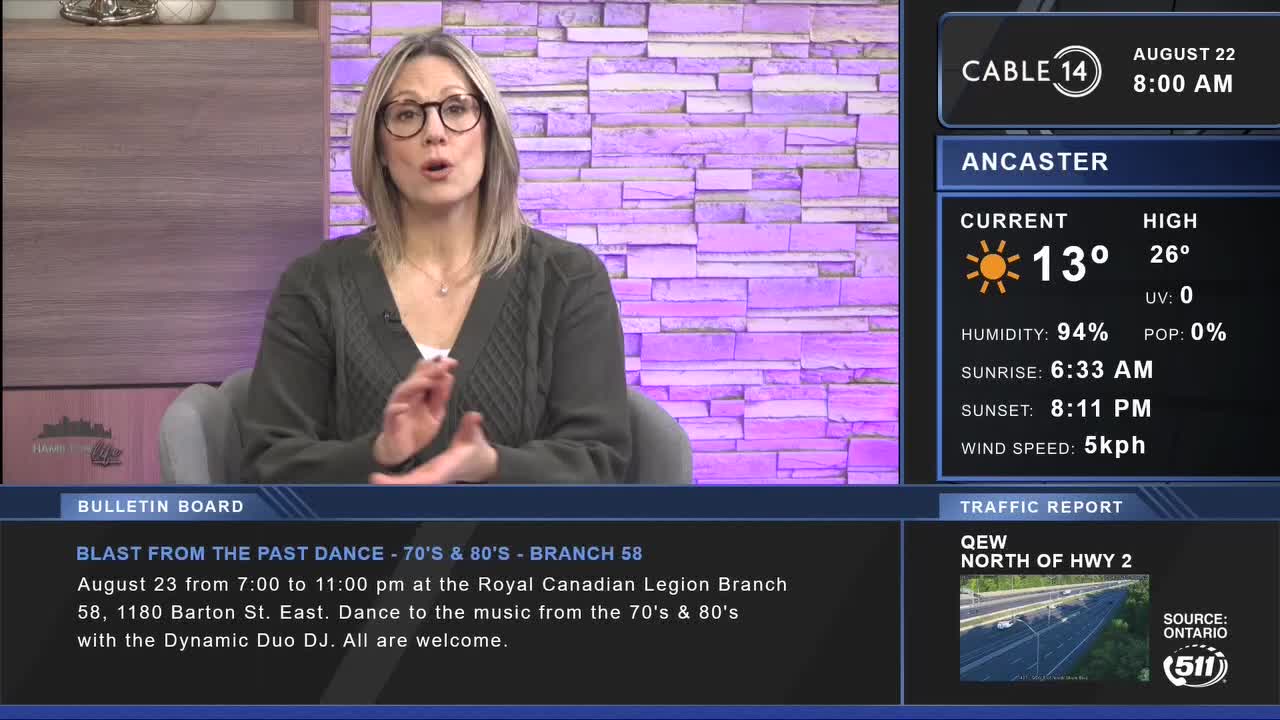 A Cable 14 Hamilton news anchor, wearing glasses and a green sweater, gestures with her hands as she speaks. The screen displays the weather in Ancaster, Ontario, and a bulletin about a "Blast from the Past" dance.

