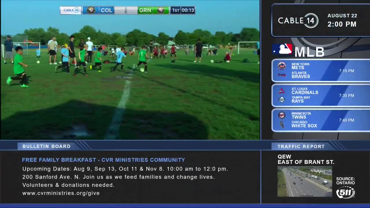 Kids in green and blue jerseys chase a soccer ball across the field, while a Cable 14 graphic in the corner shows the time and date.  The broadcast also includes a bulletin about a free family breakfast in Hamilton.
