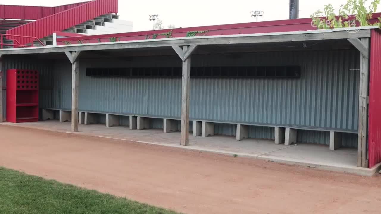 The empty dugout, with its corrugated metal siding and concrete benches, sits quiet. Overhead, the red stadium seating rises, and the Cable 14 Hamilton logo is visible on the roof.
