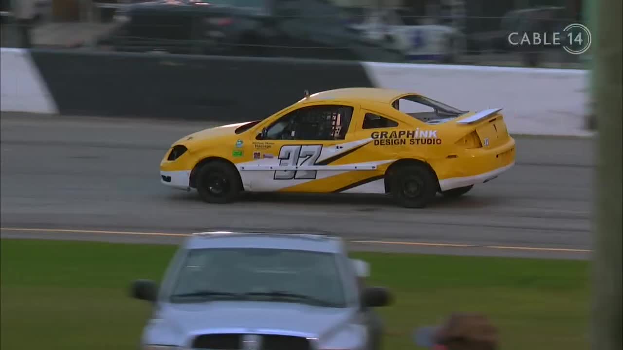 The yellow race car, number 97, is rounding the track, its white and black accents catching the light. Cable 14 Hamilton is broadcasting the race live from Canada.
