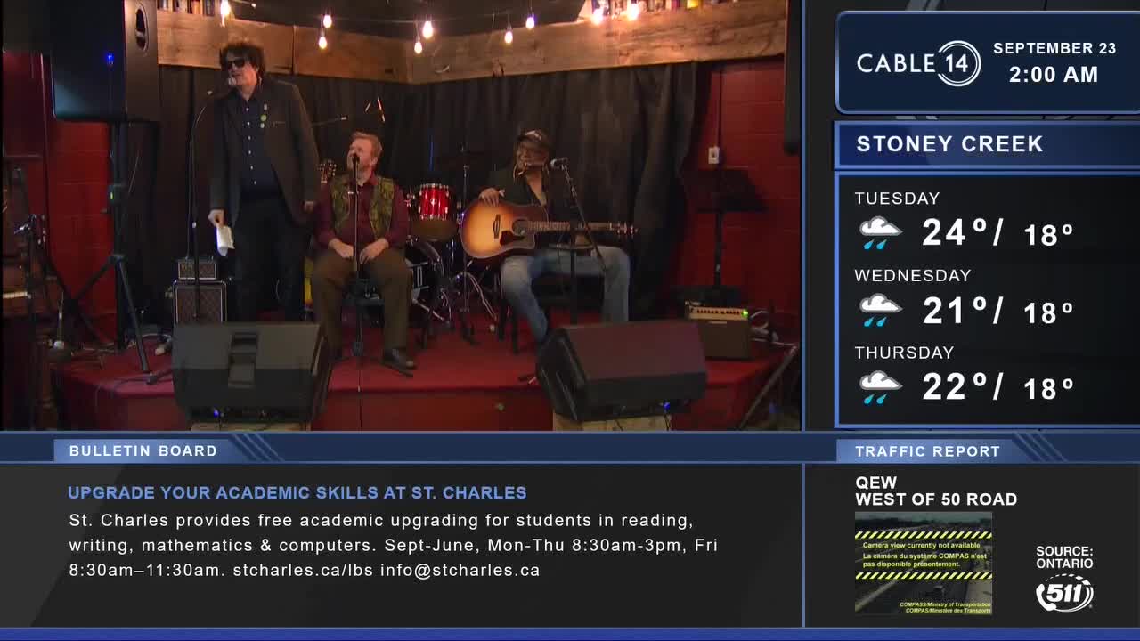 A three-piece band is set up on a small stage, lit by warm overhead bulbs. The Cable 14 Hamilton broadcast shows a musician strumming an acoustic guitar, and another is seated with a microphone.
