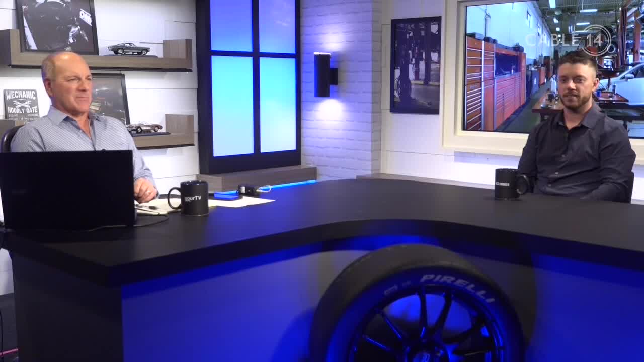 Two men sit behind a curved desk, each with a black mug in front of them. The set, lit with blue accents, has a tire embedded in the front, and a window in the background displays "Cable 14" on the screen.
