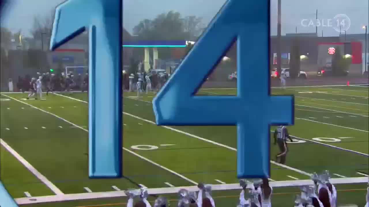 The Cable 14 logo is in the corner, and the field is wet. Players in white uniforms are running down the field, with a referee in black and white stripes following the action.
The Cable 14 logo is in the corner, and the field is wet. Players in white uniforms are running down the field, with a referee in black and white stripes following the action.