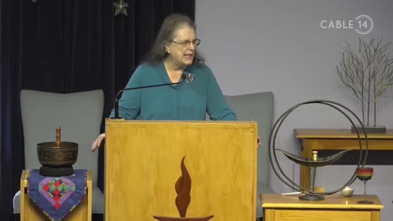 A woman in a teal top speaks from behind a wooden podium, the Cable 14 logo visible in the corner. Behind her, a simple stage is set with a singing bowl and a decorative metal sculpture.
