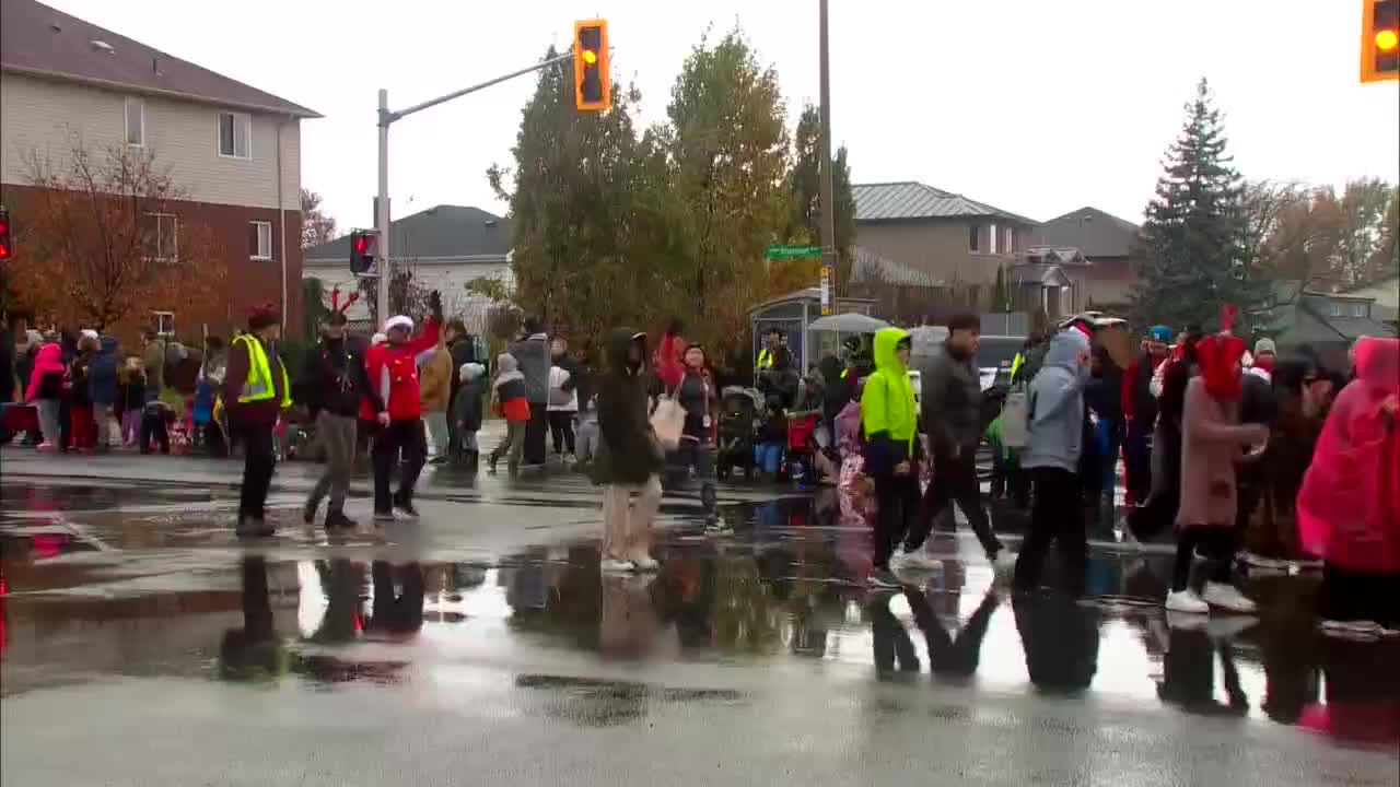 The wet street reflects the traffic lights as people cross the intersection. A Cable 14 Hamilton camera crew films the parade from the side, capturing the crowd in their rain gear.
