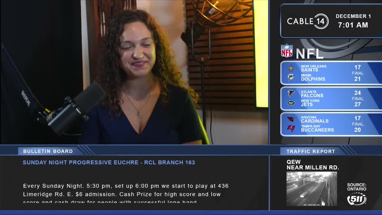 The Cable 14 Hamilton host is smiling, looking towards the camera. Behind her, a screen displays NFL scores and a traffic report from Ontario.
