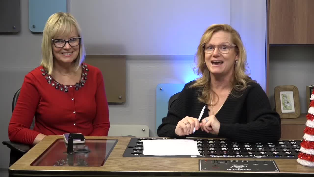 In a studio setting, two women sit behind a desk, the one on the right holding a pen and gesturing. A small, decorative Christmas tree sits on the right side of the desk.
