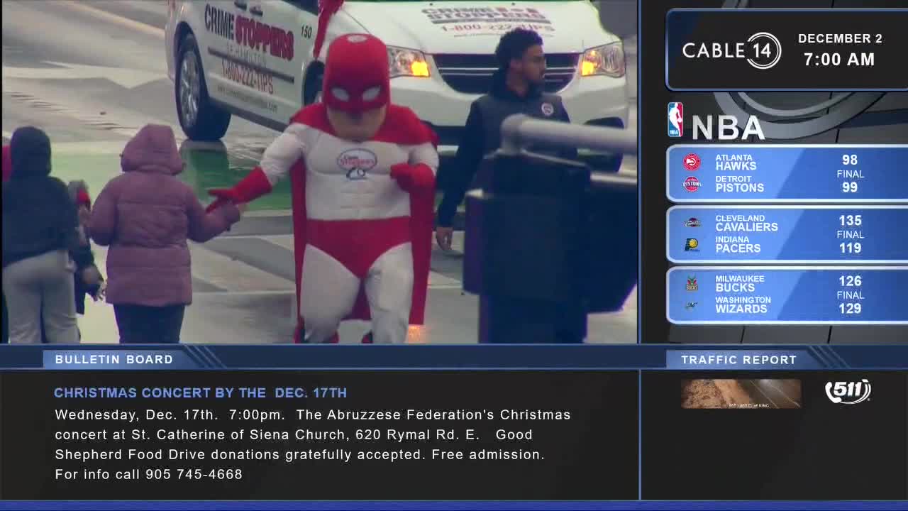 A costumed superhero, red and white, strides across a crosswalk, leading two people in winter coats. Behind him, a Crime Stoppers van idles, while Cable 14 Hamilton displays the NBA scores.
