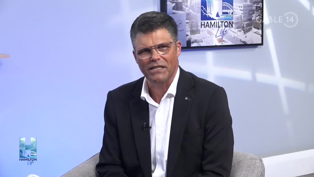 A man in a suit jacket and glasses is speaking on the Hamilton Life show on Cable 14. Behind him, the Hamilton Life logo and a cityscape scene are displayed on a screen.
