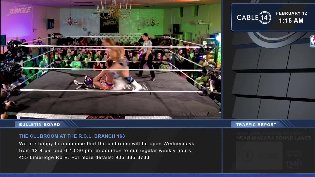 A wrestler in orange and purple shorts is pinned to the mat while another in blue and white shorts stands over him. A referee in a striped shirt watches closely as the crowd cheers.