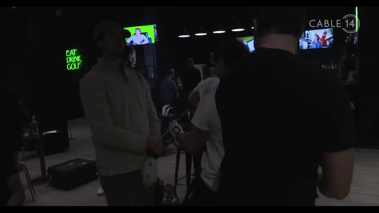 A man in a light-colored sweater stands near a neon sign that reads "EAT DRINK GOLF." Another person, wearing a black t-shirt, turns away from the camera.