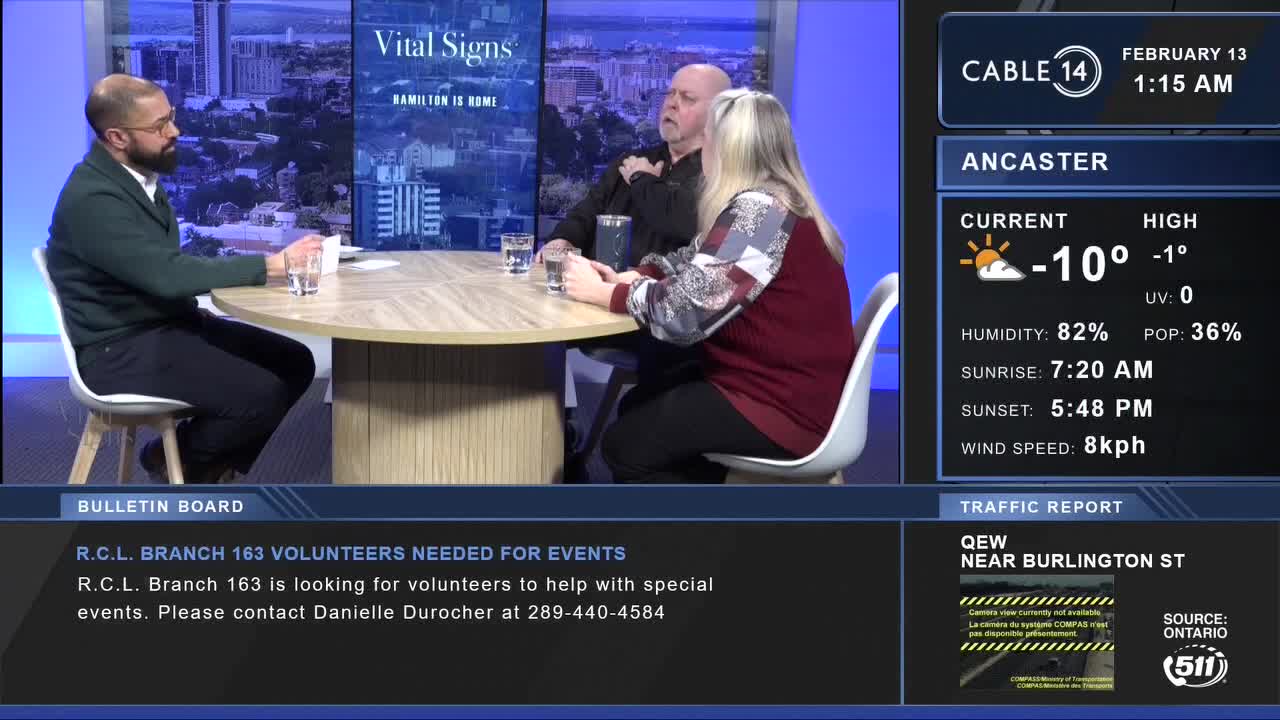 A man in a green sweater and a woman in a patterned top are seated at a round table. The Cable 14 Hamilton screen displays the date and time, along with local weather and traffic information.