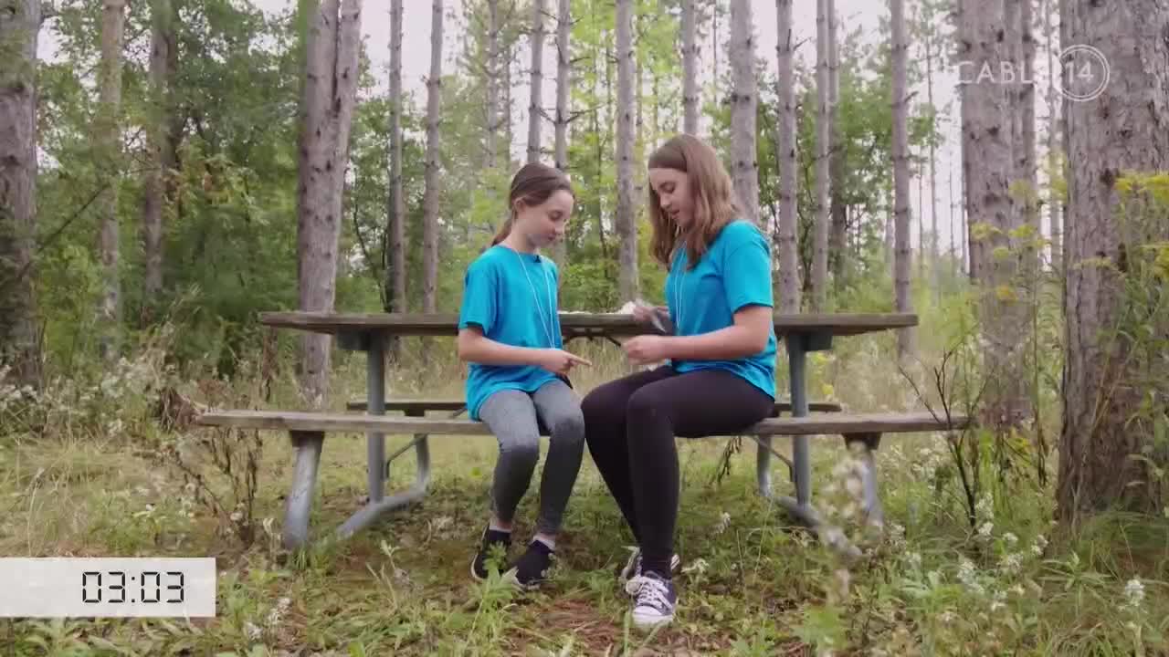 Two girls in matching blue shirts sit on a picnic bench in a Canadian forest. They are looking down at something small in their hands.