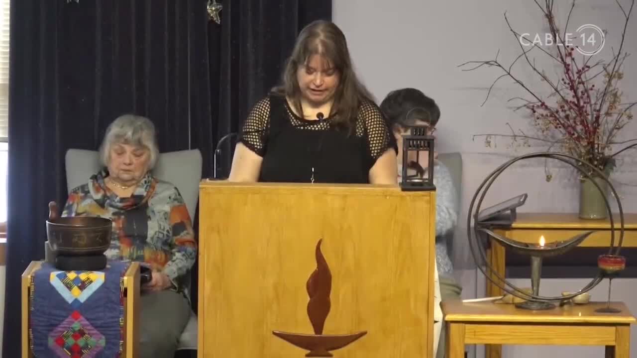 A woman stands at a wooden podium, speaking into a microphone. To her left, another woman sits in a chair, her gaze lowered towards a singing bowl. A small flame flickers in a decorative holder to the right.