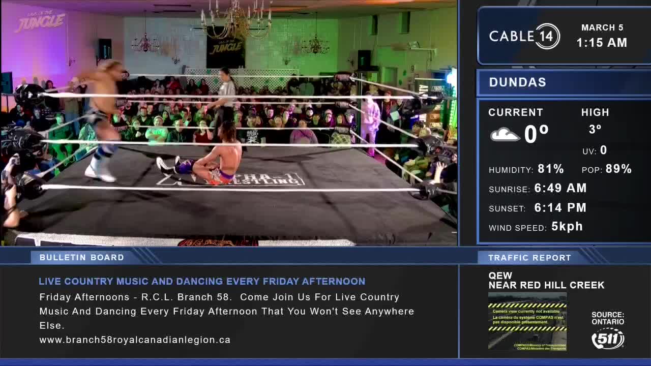 A wrestler in blue and white trunks stands over an opponent who is down on the mat. The Cable 14 Hamilton broadcast shows the match in progress, with weather and traffic updates on the side.