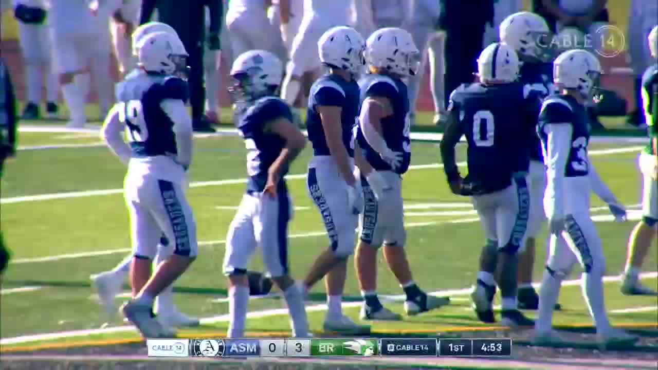 A line of football players in navy and white uniforms walks across the field. The scoreboard indicates a game is underway, broadcast by Cable 14 Hamilton.