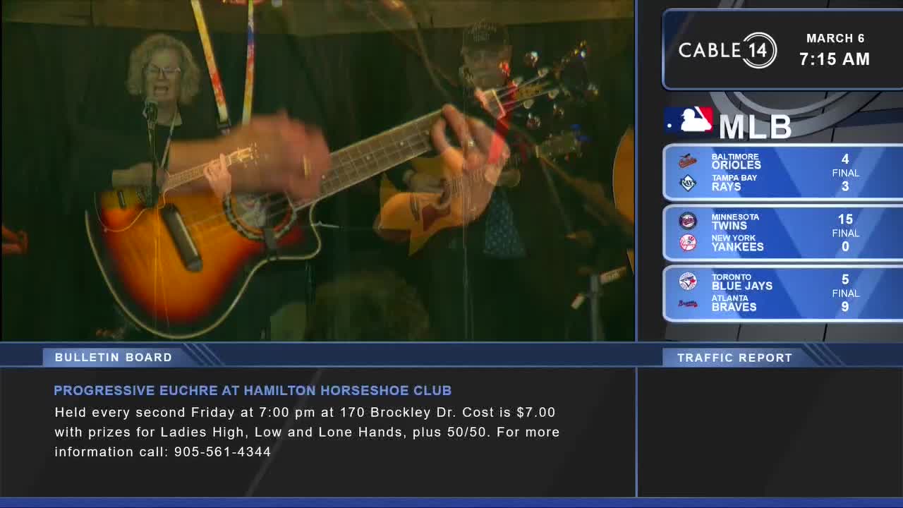 A woman sings into a microphone while a man plays an acoustic guitar. The Cable 14 Hamilton screen displays MLB scores and a bulletin about a euchre game.