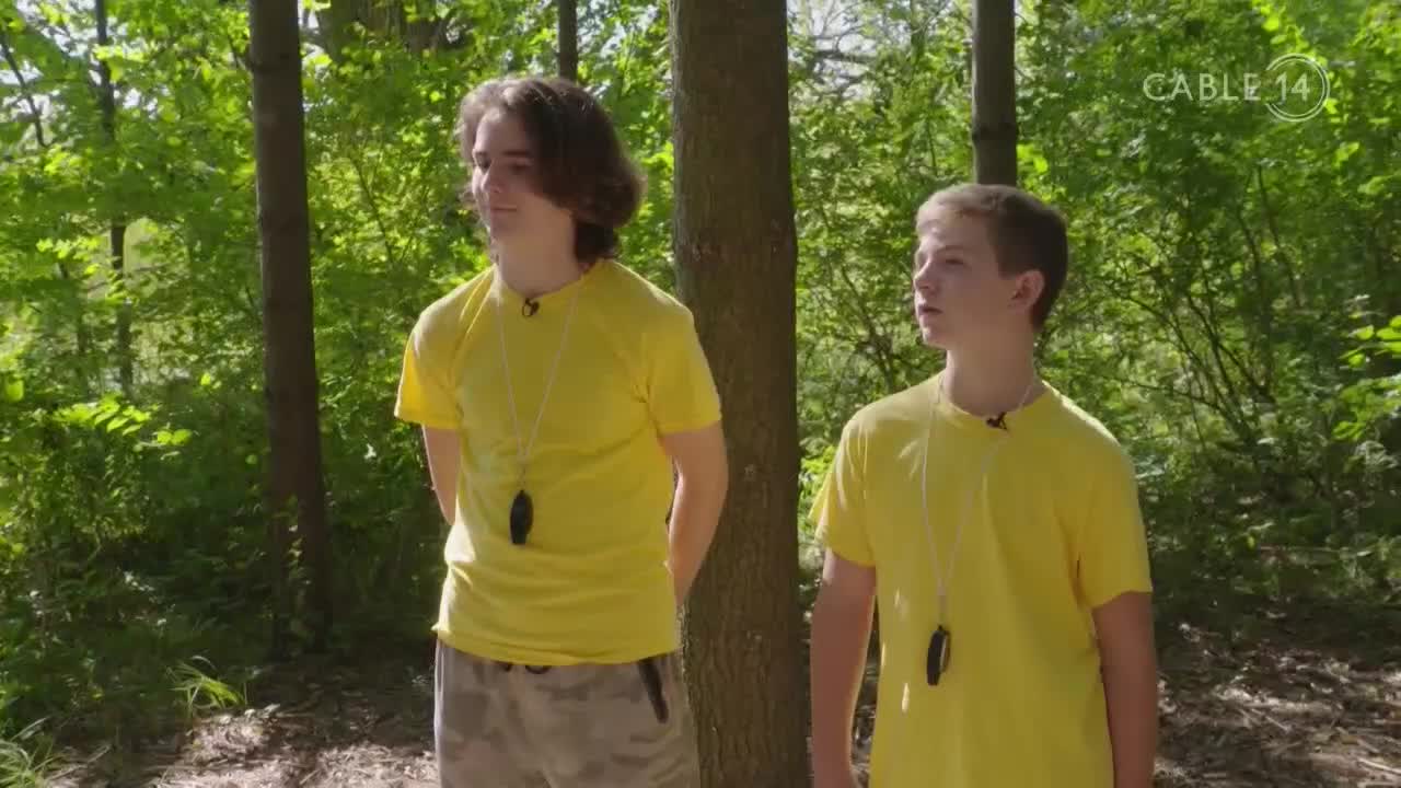 Two young men in bright yellow shirts stand in a sun-dappled forest. A Cable 14 Hamilton logo appears in the upper right corner.