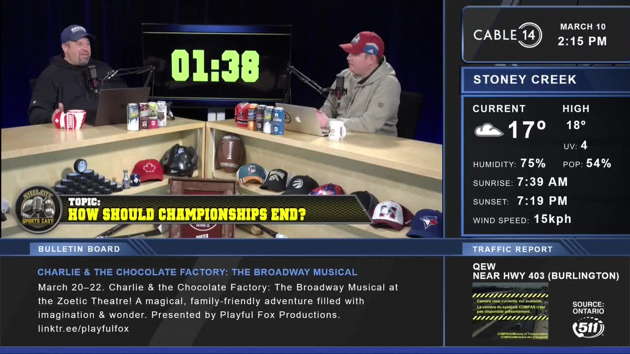 Two men are sitting at a desk, discussing something. A digital clock on the wall behind them reads 01:38. A screen to the right displays the Cable 14 Hamilton logo and local weather information for Stoney Creek.