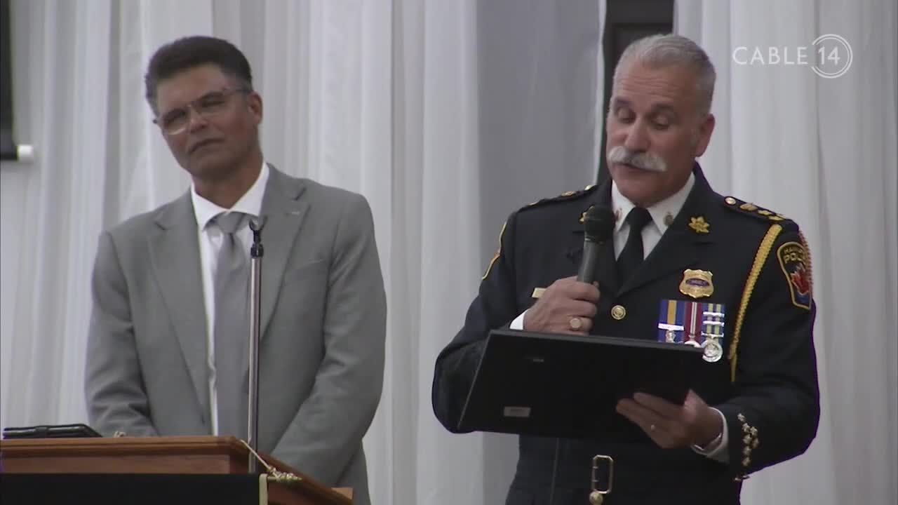A man in a police uniform with medals pinned to his chest speaks into a microphone, holding a dark plaque. Beside him, a man in a grey suit stands at a podium. The Cable 14 Hamilton logo appears in the upper right corner.