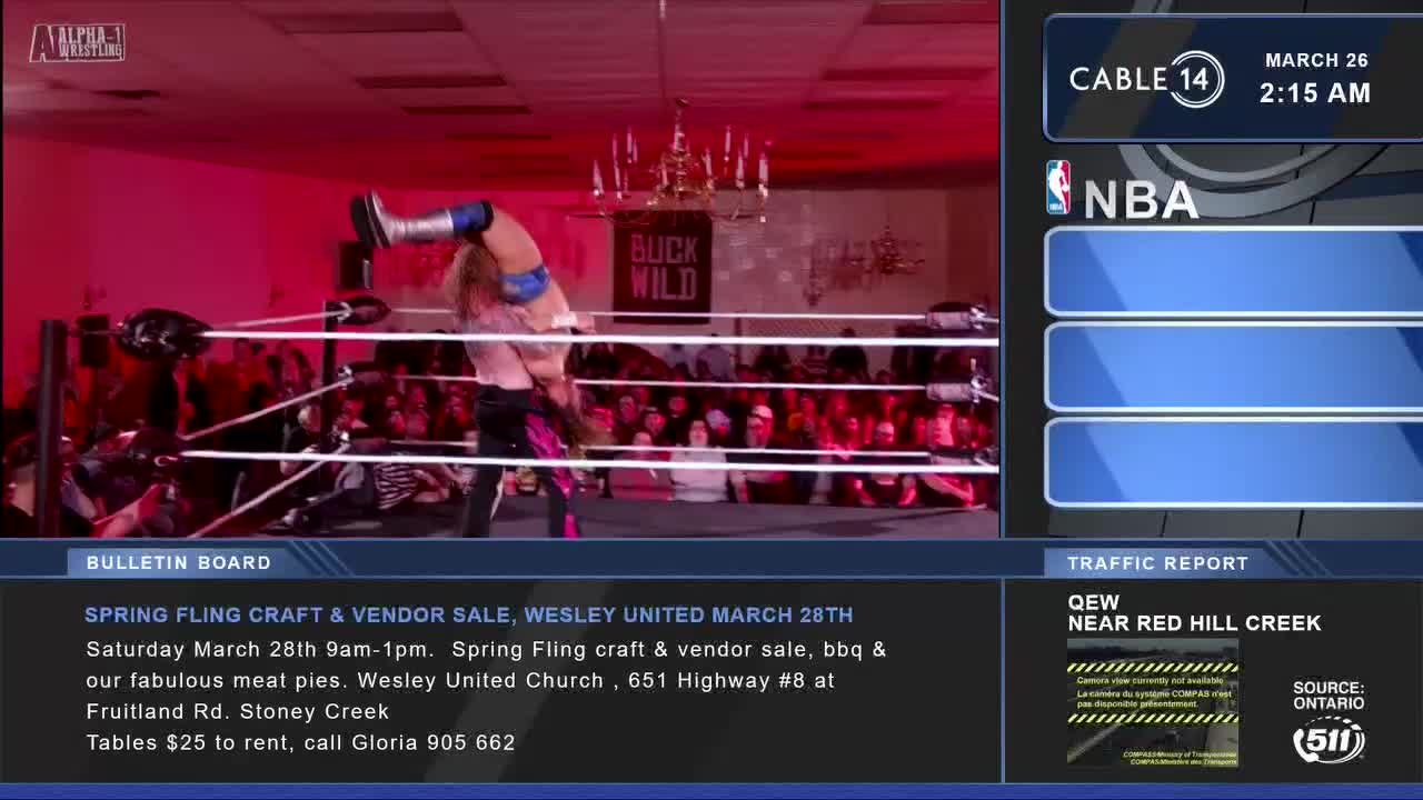 A wrestler in blue and silver is lifted upside down by another wrestler in pink and black. The crowd behind the ring is a sea of faces bathed in red light.