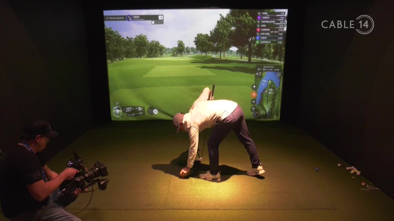 A golfer bends down to place a ball on the tee, his swing captured by a camera operator for Cable 14 Hamilton. The large screen behind them displays a virtual golf course, complete with a scorecard.