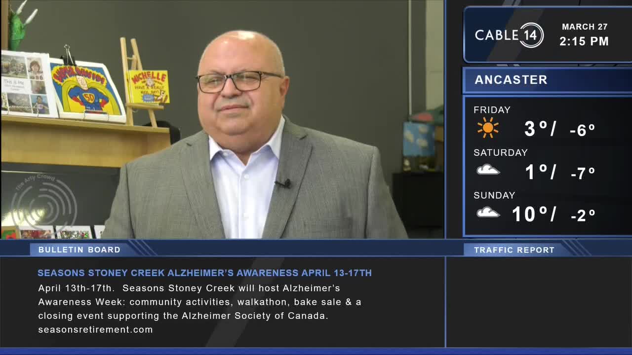 A man in a grey suit stands in front of a bookshelf, speaking into a microphone. A screen to his right displays the date, time, and weather forecast for Ancaster.