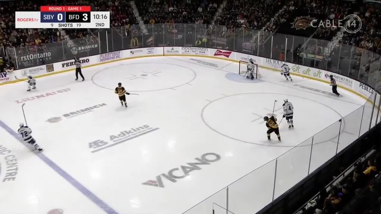 A player in a black and gold jersey skates towards the net, stick raised. Another player in white and blue is skating backwards, trying to defend.