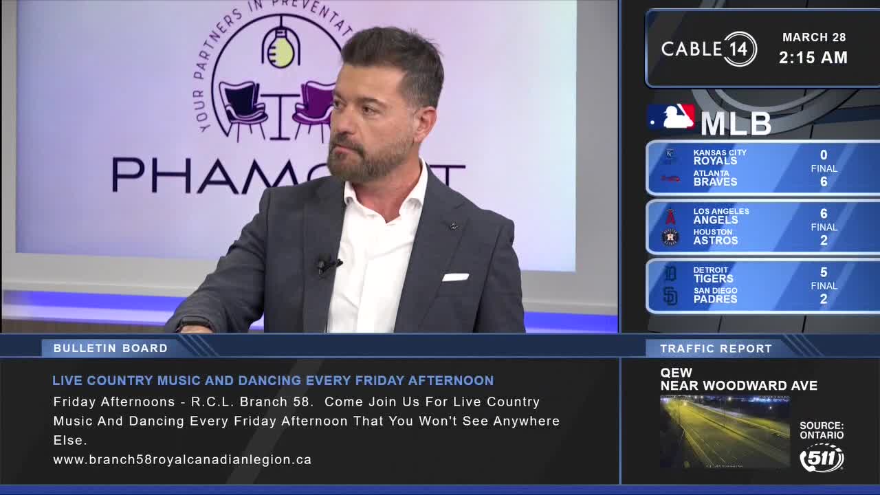 A man in a suit sits on a set, looking to his right. On the screen behind him, the Cable 14 Hamilton logo appears next to the date and time.