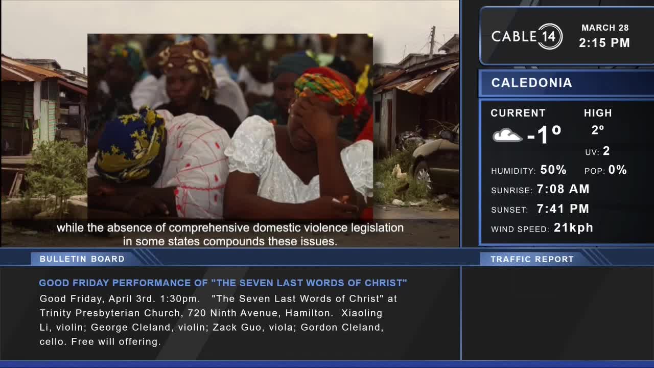 A group of people are gathered, some with heads bowed in prayer or grief. A screen displays the date and time, along with weather information for Caledonia.
