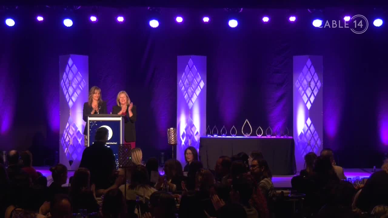 Two women stand at a podium, clapping for an award ceremony broadcast on Cable 14 Hamilton. The audience in the darkened room watches the stage, bathed in purple light.