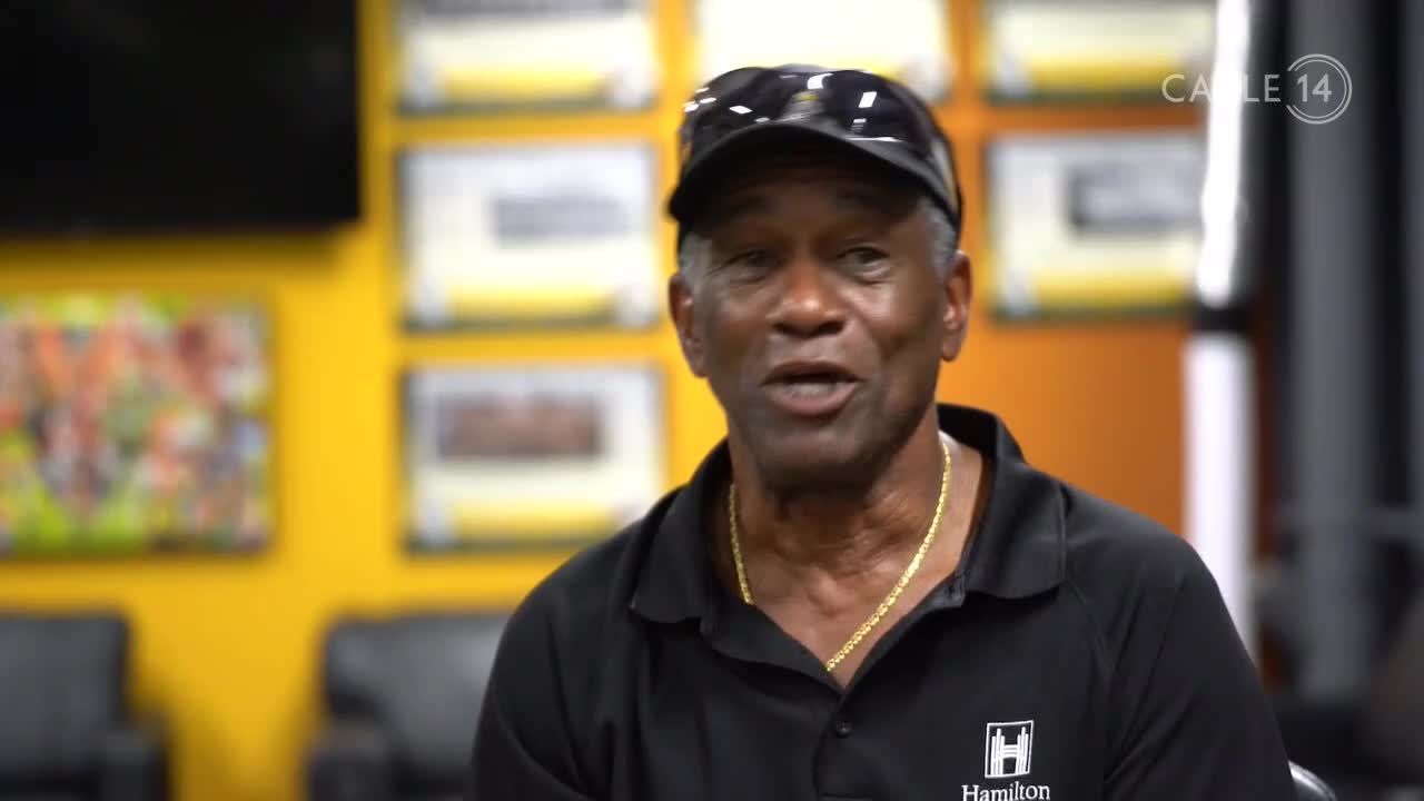 A man in a black polo shirt and baseball cap speaks directly to the camera. Behind him, the Cable 14 Hamilton logo is visible on a wall adorned with framed items.
