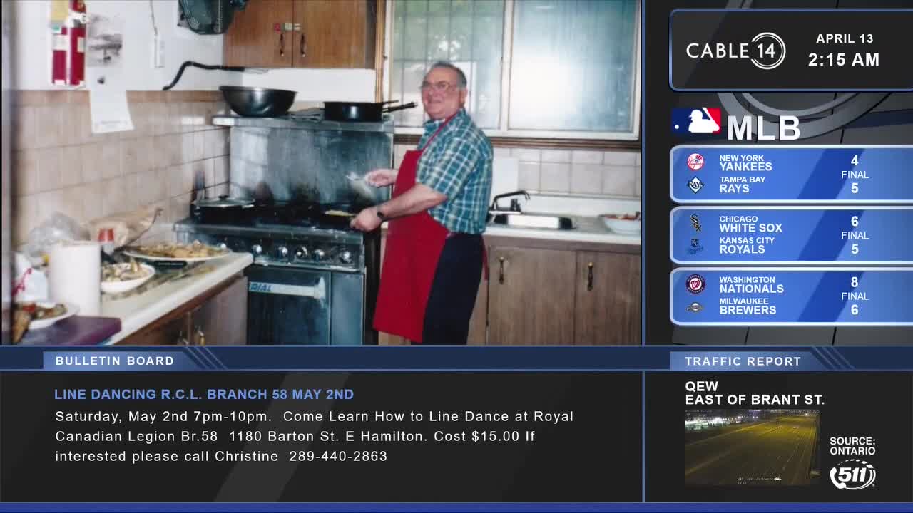 A man in a red apron is flipping something on a stovetop in a busy kitchen. On the right, Cable 14 Hamilton displays MLB scores and a traffic report for the QEW.