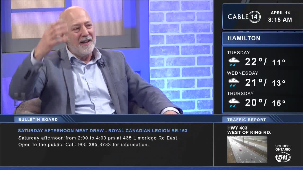 A man with a grey jacket gestures with his hand while speaking. On the screen beside him, Cable 14 Hamilton displays the day's weather and traffic updates.