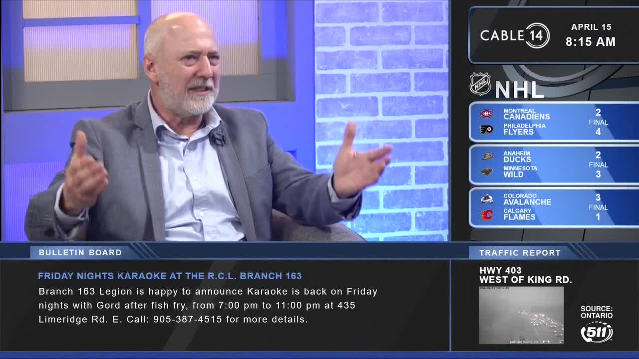 A man in a grey suit gestures emphatically while speaking. On the screen beside him, Cable 14 Hamilton displays NHL scores and a traffic report for Highway 403.