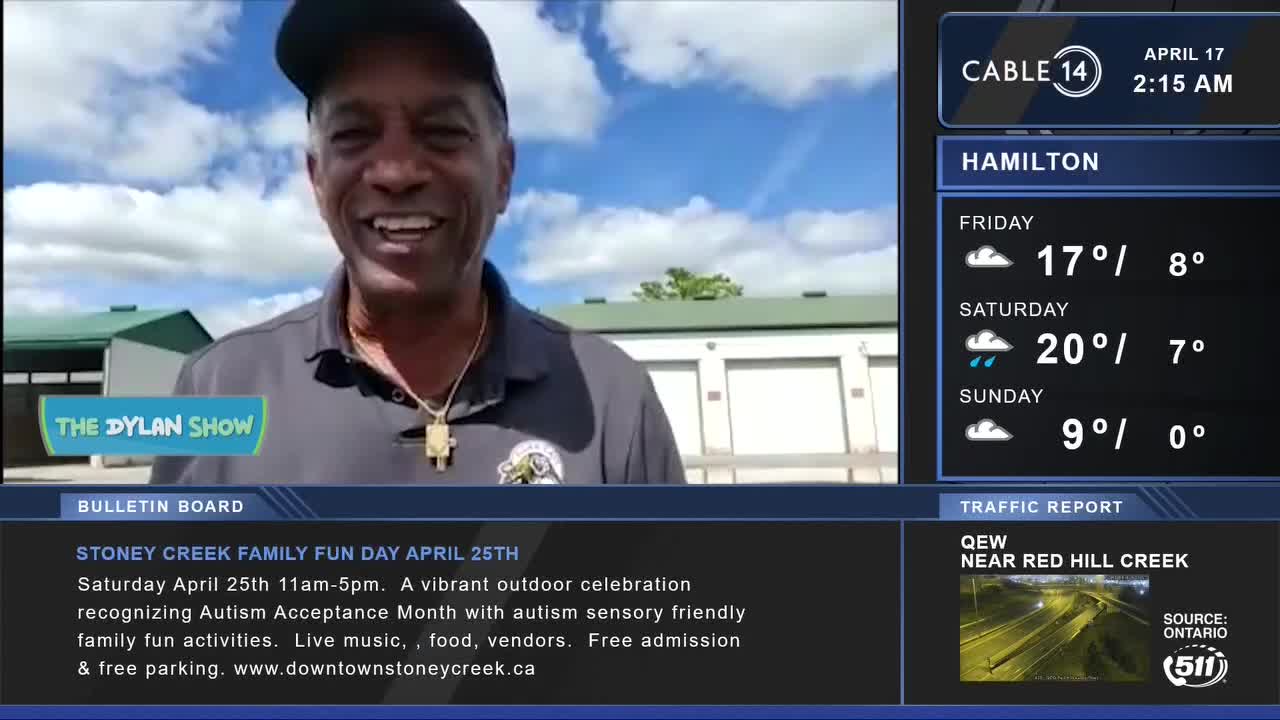 A man smiles widely, wearing a grey polo shirt and a gold chain. Behind him, a bright blue sky with fluffy clouds is visible above a row of beige storage units.