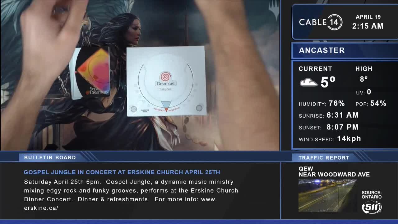 A hand moves a white Sega Dreamcast console and a game disc across a dark, patterned surface. Cable 14 Hamilton displays the current time and weather for Ancaster.