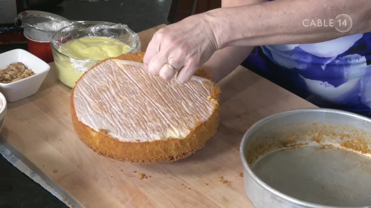 Fingers peel parchment paper from a cake layer, revealing a creamy filling. This moment, captured for Cable 14 Hamilton, shows the careful assembly of a dessert.