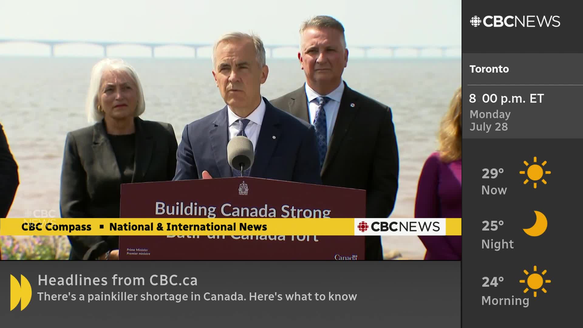 A man in a suit speaks into a microphone, standing behind a podium that reads "Building Canada Strong."  Behind him, a bridge stretches across the water under a bright sky, as reported by CBC News Network.
