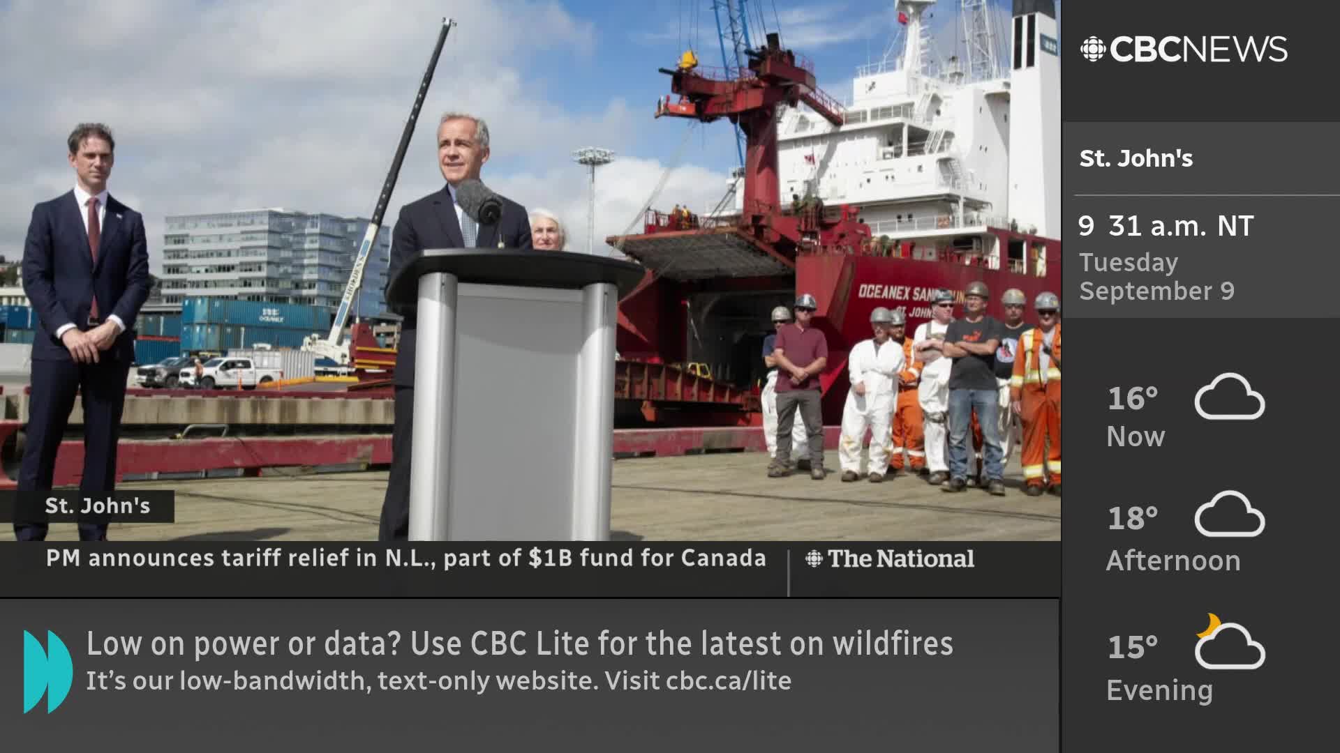 A man in a suit speaks from behind a podium, with a large red and white ship looming behind him. A group of workers in hard hats and coveralls stand to the side, as a CBC News Network graphic displays the weather in St. John's.
