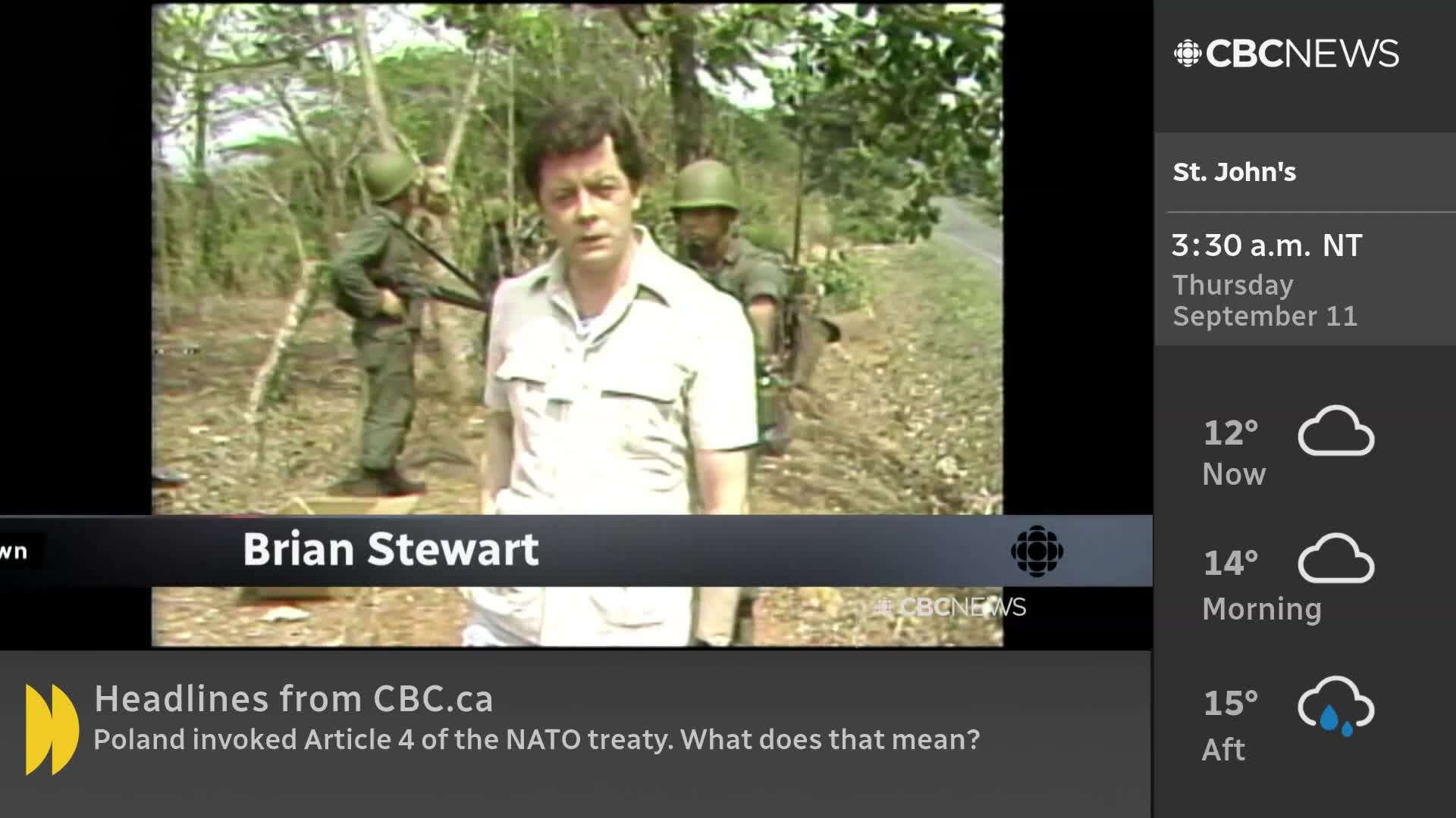 Brian Stewart, a CBC News reporter, stands in front of the camera, speaking. Behind him, two soldiers in green uniforms are visible, one holding a rifle.
