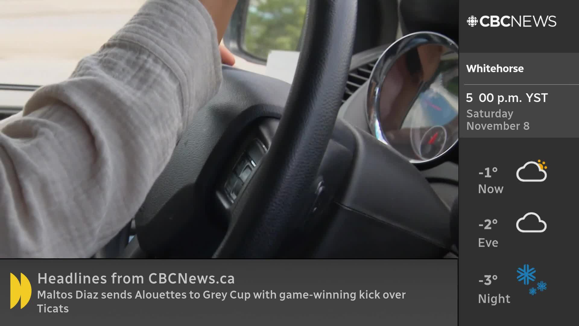 A hand rests on the steering wheel, the sleeve of a light shirt visible. CBC News Network's Whitehorse weather report is displayed on the screen.
