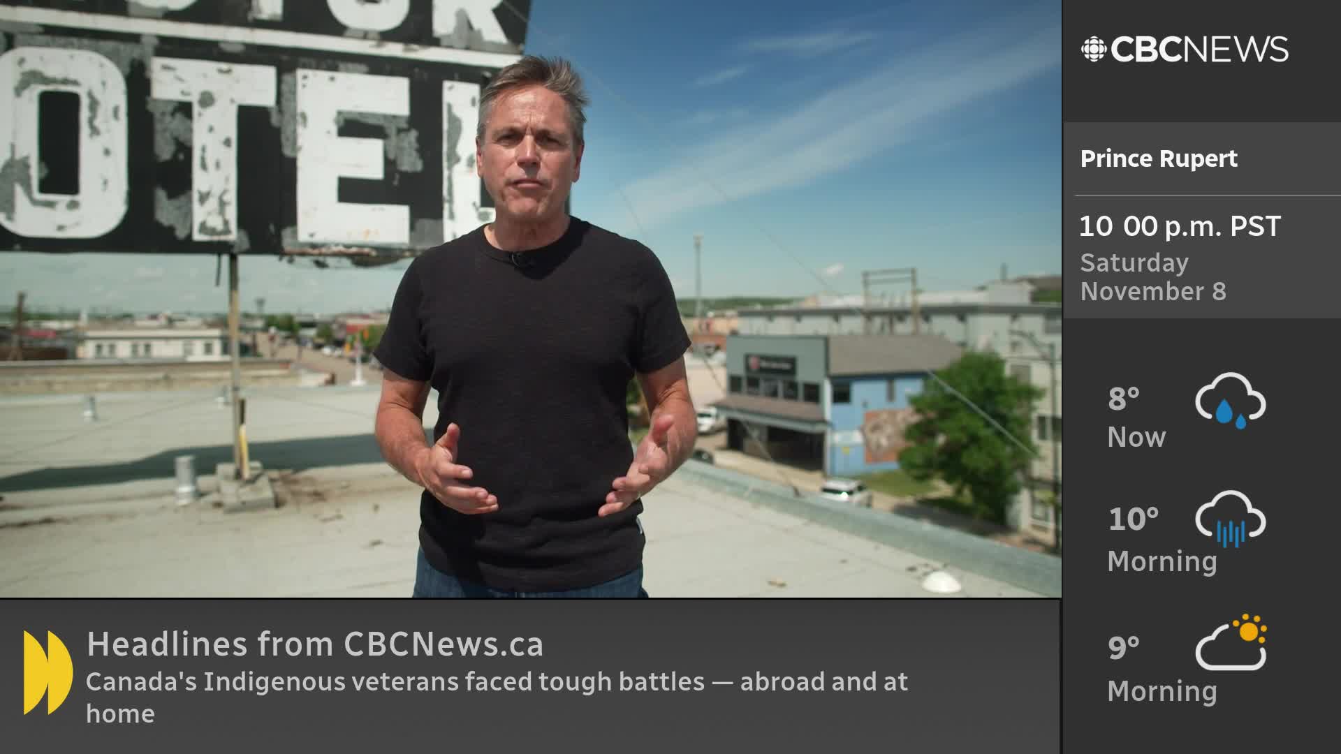 A man in a black shirt stands on a rooftop, gesturing with his hands as he speaks. Behind him, a weathered "MOTOR HOTEL" sign looms against a bright blue sky, the CBC News Network logo visible in the corner.
