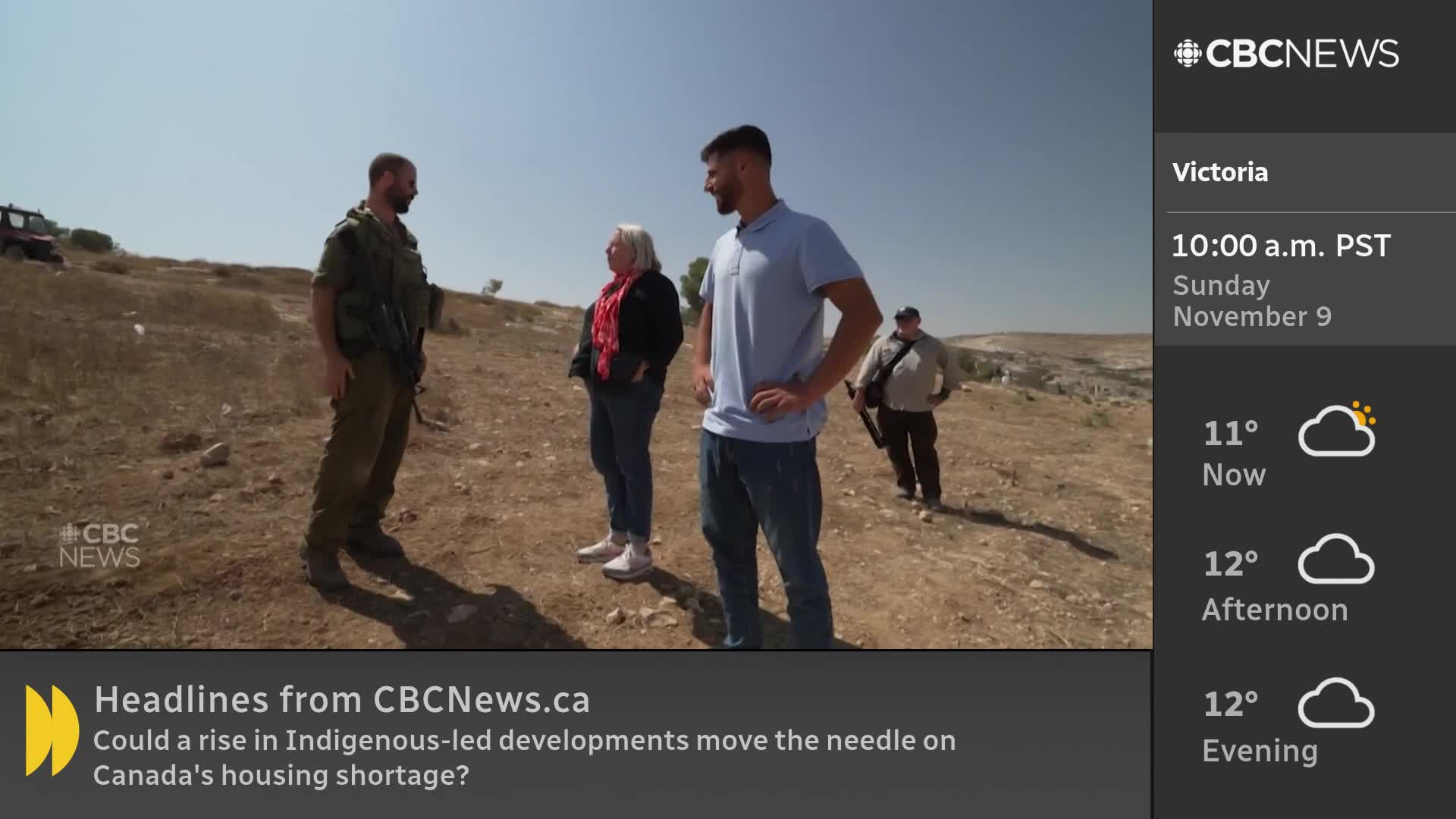 A group of people stand on a dry, hilly landscape; one is dressed in military fatigues and holding a rifle. CBC News Network's logo is visible in the corner, and the weather forecast for Victoria, Canada, is displayed alongside the image.

