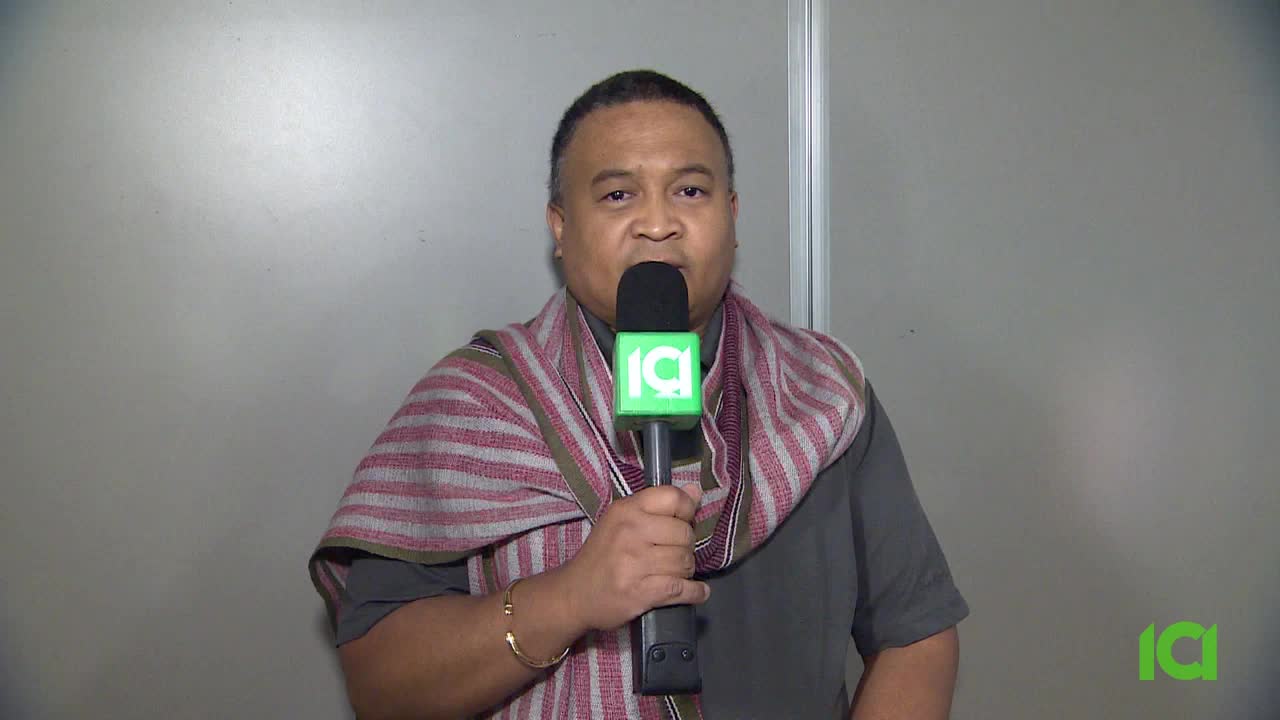 A man in a gray shirt and patterned scarf holds a microphone with a green logo. He's facing the camera, speaking directly, likely for CFHD-DT in Canada.
