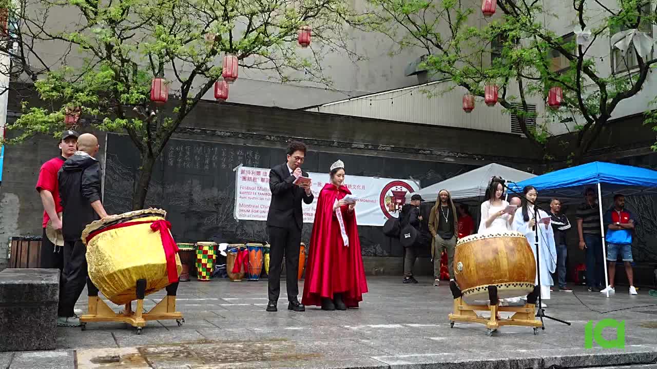 A man in a suit and a woman in a red cape are speaking into microphones on a stage, reading from papers. Two large drums sit on either side, ready for the ceremony, as CFHD-DT likely broadcasts the event from Canada.
